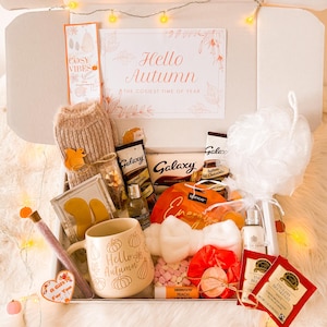 May include: An autumn-themed gift box containing a mug with pumpkins and the text "Hello Autumn", chocolates, tea bags, and a bath puff. The box also includes a knitted item, a lip balm, and a card that says "Hello Autumn". Decorated with fairy lights.