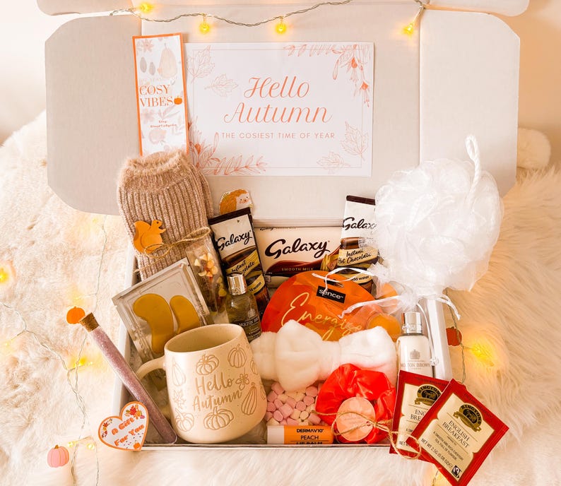 May include: An autumn-themed gift box containing a mug with pumpkins and the text "Hello Autumn", chocolates, tea bags, and a bath puff. The box also includes a knitted item, a lip balm, and a card that says "Hello Autumn". Decorated with fairy lights.