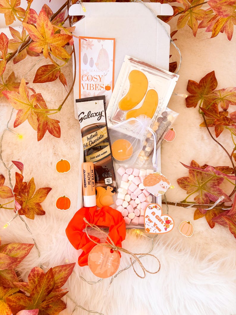 May include: A gift box containing autumn-themed treats. It features Galaxy instant hot chocolate, peach lip balm, eye patches, marshmallows, and an orange scrunchie. The box is decorated with fall leaves and the text "The Season of Cosy Vibes".