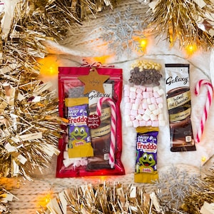 May include: A festive gift set featuring Galaxy instant hot chocolate, Dairy Milk Freddo chocolate bars, marshmallows, and chocolate chips. The set includes a red bag with a star-shaped tag and a candy cane, all surrounded by gold tinsel.