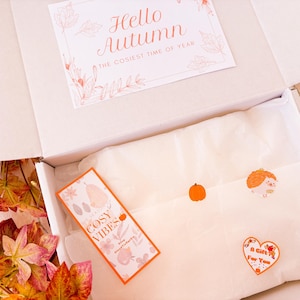 May include: A white box, open to reveal a card saying "Hello Autumn" and a "Cosy Vibes" tag. Inside, tissue paper is decorated with stickers of a pumpkin, hedgehog, and heart. Autumn leaves and a knitted blanket are visible.
