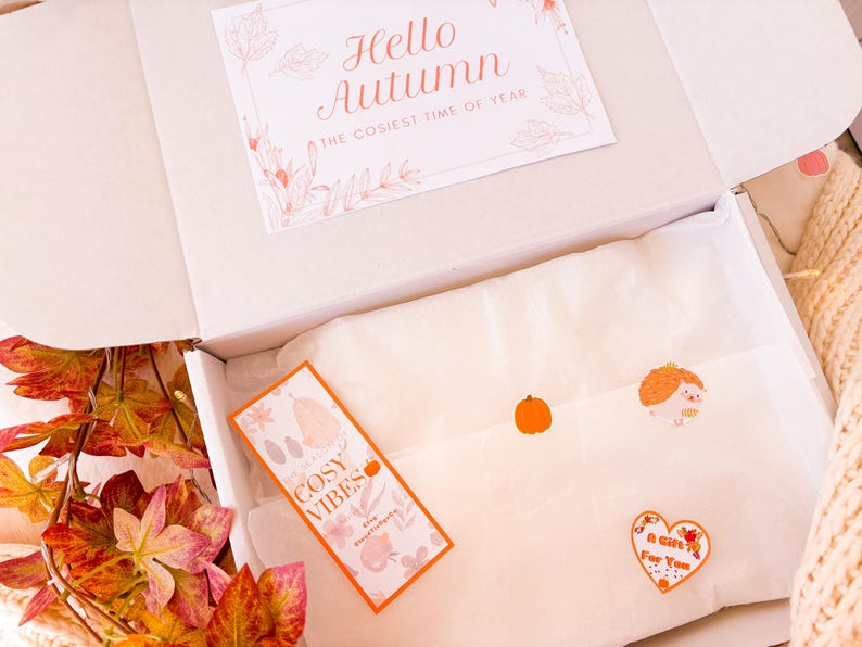 May include: A white box, open to reveal a card saying "Hello Autumn" and a "Cosy Vibes" tag. Inside, tissue paper is decorated with stickers of a pumpkin, hedgehog, and heart. Autumn leaves and a knitted blanket are visible.