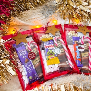 May include: Three red cellophane gift bags filled with candy bars, marshmallows, and candy canes. Each bag is topped with a star-shaped tag. The bags are surrounded by gold tinsel and a white knitted blanket.