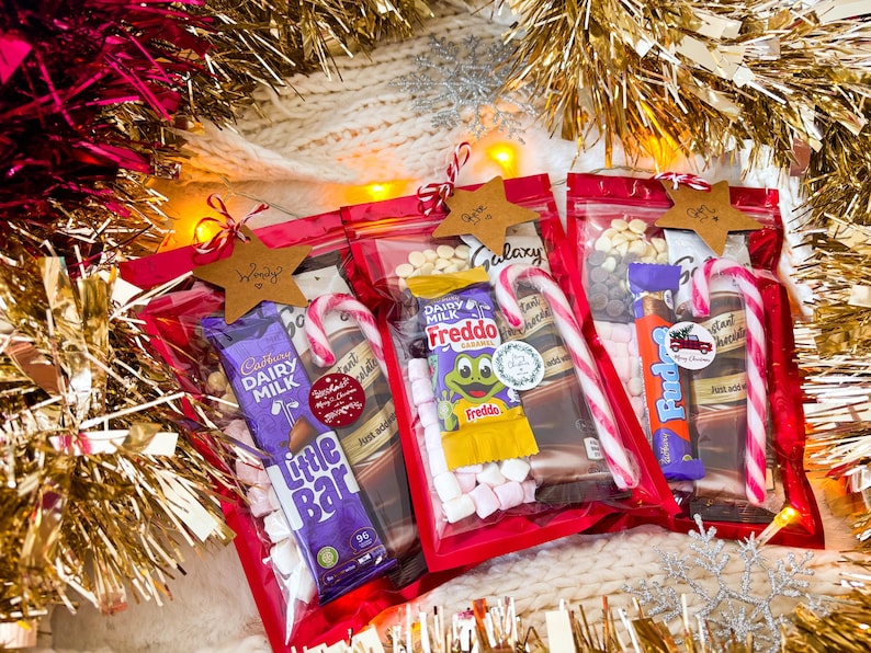 May include: Three red cellophane gift bags filled with candy bars, marshmallows, and candy canes. Each bag is topped with a star-shaped tag. The bags are surrounded by gold tinsel and a white knitted blanket.