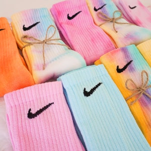 May include: A collection of colorful tie-dye socks, each featuring the Nike swoosh logo in black. The socks are in shades of orange, pink, blue, yellow, and tie-dye patterns, some tied with twine. These socks are a stylish accessory.