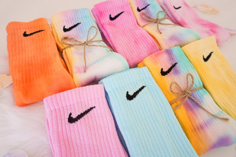 May include: A collection of colorful tie-dye socks, each featuring the Nike swoosh logo in black. The socks are in shades of orange, pink, blue, yellow, and tie-dye patterns, some tied with twine. These socks are a stylish accessory.