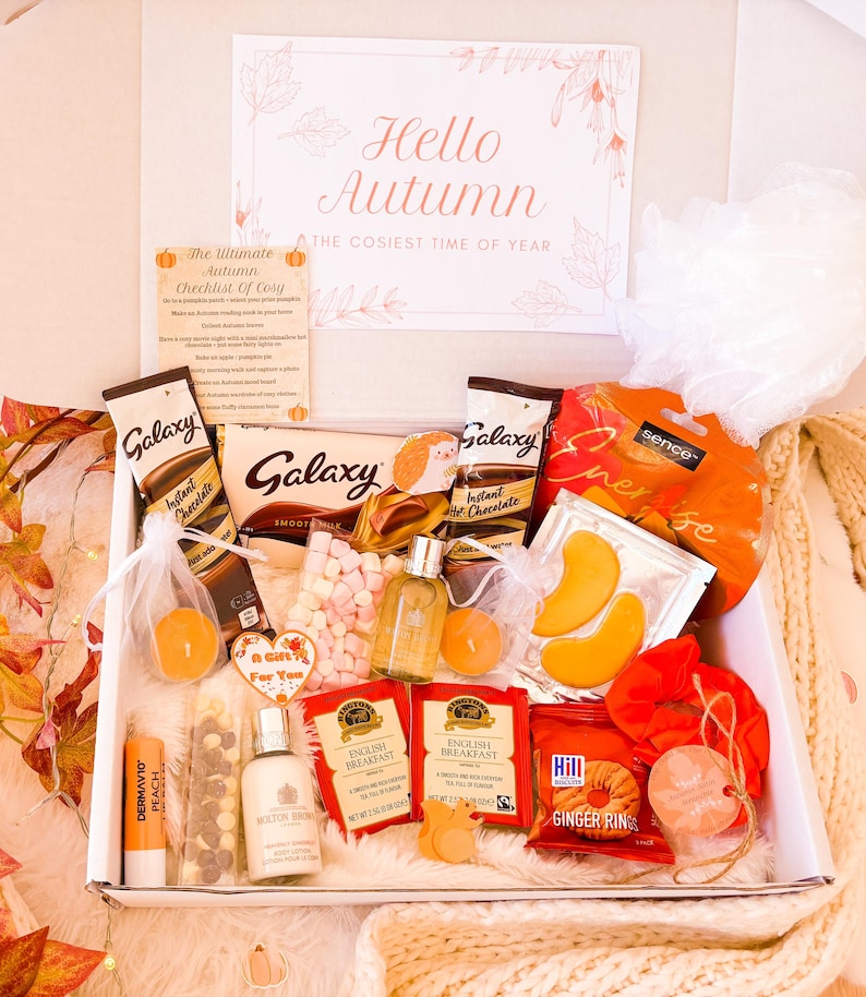 May include: A gift box with autumn-themed items, including Galaxy chocolate, instant hot chocolate sachets, English Breakfast tea bags, and ginger rings. The box is decorated with a card that reads "Hello Autumn, The Cosiest Time of Year".