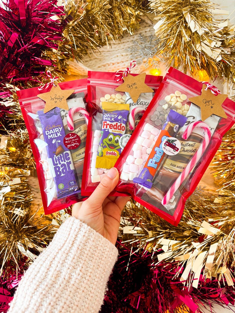 May include: Four red zipper bags filled with candy and treats, including chocolate bars, marshmallows, and candy canes. Each bag is adorned with a star-shaped tag. The background features gold and red tinsel.