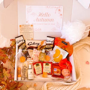 May include: A gift box with autumn-themed treats and self-care items. Inside are chocolate, tea, marshmallows, and bath products. A card reads "Hello Autumn". The color palette is warm, with orange, brown, and white tones, evoking a cozy atmosphere.