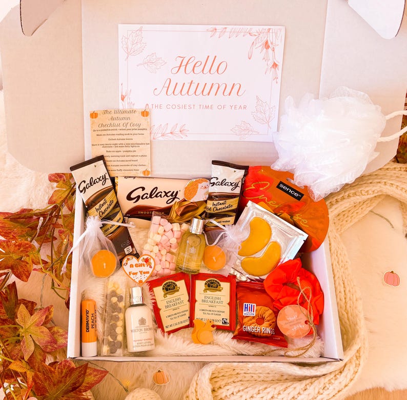 May include: A gift box with autumn-themed treats and self-care items. Inside are chocolate, tea, marshmallows, and bath products. A card reads "Hello Autumn". The color palette is warm, with orange, brown, and white tones, evoking a cozy atmosphere.