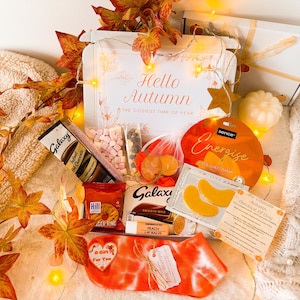 May include: An autumn-themed gift box with the text "Hello Autumn". The box contains Galaxy hot chocolate, marshmallows, a peach lip balm, and other treats. Orange and white socks are also included, along with decorative fall leaves.