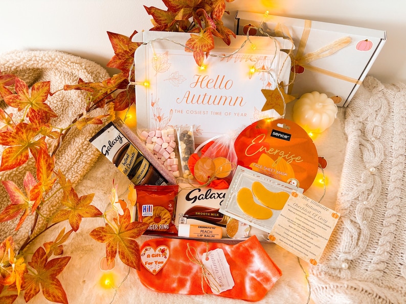 May include: An autumn-themed gift box with the text "Hello Autumn". The box contains Galaxy hot chocolate, marshmallows, a peach lip balm, and other treats. Orange and white socks are also included, along with decorative fall leaves.