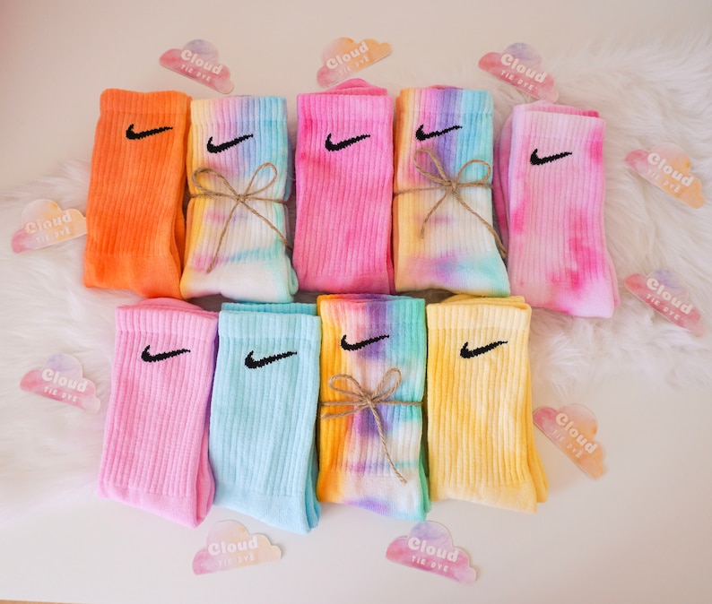 May include: A collection of colorful tie-dye socks with the Nike swoosh logo embroidered in black. The socks are in various shades of pink, orange, yellow, and blue. Some pairs are tied with twine. The image includes cloud-shaped stickers that say "Cloud Tie Dye".
