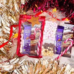 May include: A festive gift set with hot chocolate ingredients. Includes Cadbury Dairy Milk Little Bar chocolate, marshmallows, chocolate chips, and candy canes, all arranged with gold and red tinsel.