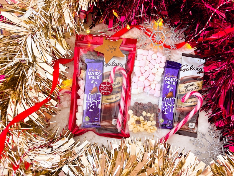 May include: A festive gift set with hot chocolate ingredients. Includes Cadbury Dairy Milk Little Bar chocolate, marshmallows, chocolate chips, and candy canes, all arranged with gold and red tinsel.
