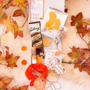 May include: A gift box filled with fall-themed items. Includes a packet of Galaxy instant hot chocolate, peach lip balm, eye patches, marshmallows, and a scrunchie. The box is decorated with autumn leaves and pumpkin stickers. The text "The Season of Cosy Vibes" is visible.