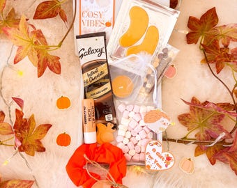 May include: A gift box filled with fall-themed items. Includes a packet of Galaxy instant hot chocolate, peach lip balm, eye patches, marshmallows, and a scrunchie. The box is decorated with autumn leaves and pumpkin stickers. The text "The Season of Cosy Vibes" is visible.