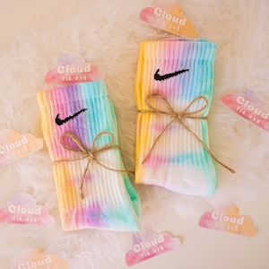 May include: Two pairs of tie-dye socks with the Nike swoosh logo, tied with twine. The socks feature a pastel rainbow design of yellow, pink, purple, and blue. Cloud-shaped tags with the text "Cloud Tie Dye" are scattered around the socks.