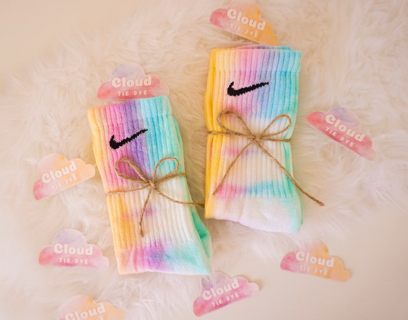 May include: Two pairs of tie-dye socks with the Nike swoosh logo, tied with twine. The socks feature a pastel rainbow design of yellow, pink, purple, and blue. Cloud-shaped tags with the text "Cloud Tie Dye" are scattered around the socks.