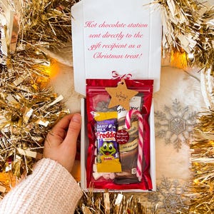 May include: A gift box with a red-sealed bag filled with hot chocolate ingredients, including a candy cane, chocolate bar, and marshmallows. The box is decorated with a star-shaped tag and the text "Hot chocolate station sent directly to the gift recipient as a Christmas treat!"