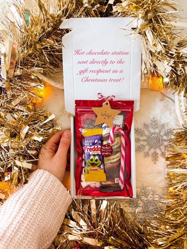 May include: A gift box with a red-sealed bag filled with hot chocolate ingredients, including a candy cane, chocolate bar, and marshmallows. The box is decorated with a star-shaped tag and the text "Hot chocolate station sent directly to the gift recipient as a Christmas treat!"