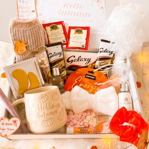 May include: An autumn-themed gift box containing a variety of items. Includes a mug with "Hello Autumn" text, tea bags, Galaxy chocolate, a bow-shaped headband, and a scrunchie. Other items include eye masks, lip balm, and a small sweater.