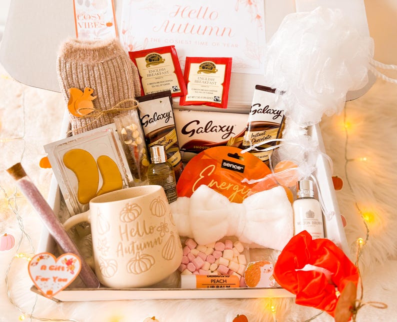 May include: An autumn-themed gift box containing a variety of items. Includes a mug with "Hello Autumn" text, tea bags, Galaxy chocolate, a bow-shaped headband, and a scrunchie. Other items include eye masks, lip balm, and a small sweater.