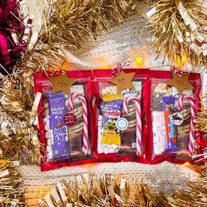 May include: Three red cellophane bags filled with candy, chocolate bars, and marshmallows, each with a candy cane. The bags are decorated with star-shaped tags and surrounded by gold and pink tinsel.