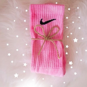 May include: A pair of pink socks with the Nike swoosh logo in black. The socks are tied with a brown twine bow. The background is white and fluffy, with white star shapes scattered around the socks.