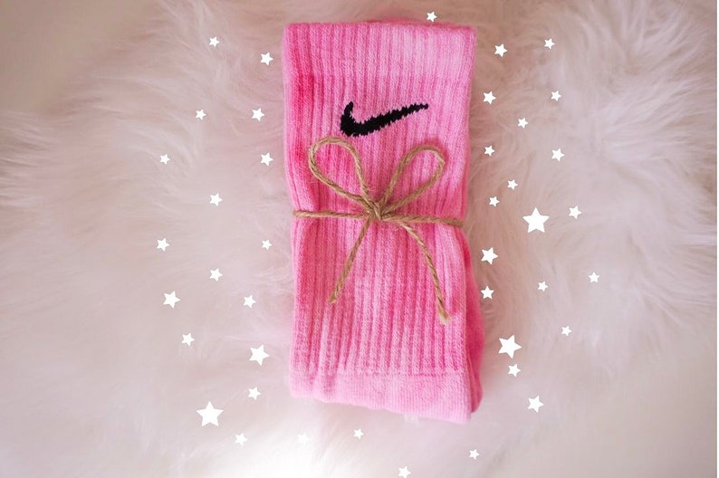 May include: A pair of pink socks with the Nike swoosh logo in black. The socks are tied with a brown twine bow. The background is white and fluffy, with white star shapes scattered around the socks.