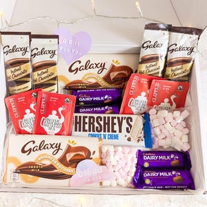 May include: A gift box filled with Galaxy instant hot chocolate, Clipper organic English breakfast tea, Hershey's Cookies 'n' Creme chocolate bar, Cadbury Dairy Milk chocolate, and marshmallows. The box includes a heart-shaped gift tag.