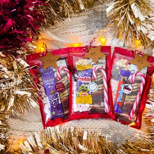 May include: Three red cellophane bags filled with candy bars, marshmallows, and chocolate chips, each adorned with a candy cane and a star-shaped tag. The bags are surrounded by gold and pink tinsel, creating a festive holiday theme.