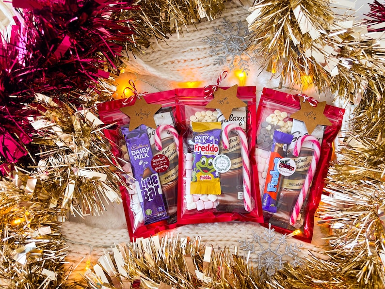 May include: Three red cellophane bags filled with candy bars, marshmallows, and chocolate chips, each adorned with a candy cane and a star-shaped tag. The bags are surrounded by gold and pink tinsel, creating a festive holiday theme.