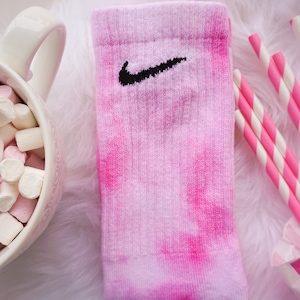 May include: Pink tie-dye sock with a black Nike swoosh logo. The sock is next to a bowl of pink and white marshmallows, pink and white striped straws, and a pink and white striped paper bag.