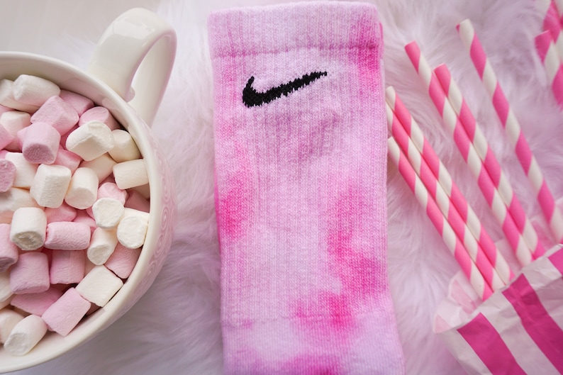 May include: Pink tie-dye sock with a black Nike swoosh logo. The sock is next to a bowl of pink and white marshmallows, pink and white striped straws, and a pink and white striped paper bag.