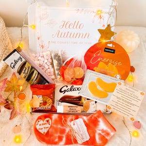 May include: An autumn-themed gift box containing Galaxy hot chocolate, a face sheet mask, Galaxy smooth milk chocolate, ginger rings, peach lip balm, and tie-dye socks. The box is decorated with fairy lights and the text "Hello Autumn: The Cosiest Time of Year".