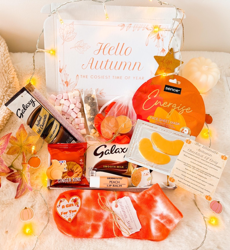 May include: An autumn-themed gift box containing Galaxy hot chocolate, a face sheet mask, Galaxy smooth milk chocolate, ginger rings, peach lip balm, and tie-dye socks. The box is decorated with fairy lights and the text "Hello Autumn: The Cosiest Time of Year".