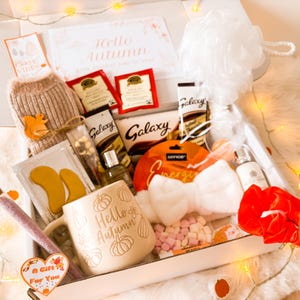 Pode incluir: Uma caixa de presente cheia de itens com tema de outono. Inclui uma caneca com o texto "Hello Autumn", saquinhos de chá, chocolate, uma esponja de banho e um pequeno suéter com um aplique de esquilo. Luzes decorativas e uma etiqueta em forma de coração "A Gift For You" também estão presentes.