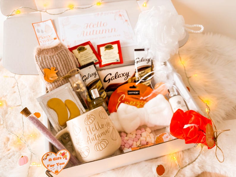 May include: A gift box containing autumn-themed items. It features a mug with "Hello Autumn" text, tea bags, chocolate, a bath pouf, and a small sweater with a squirrel applique. Decorative lights and a "A Gift For You" tag are also included.