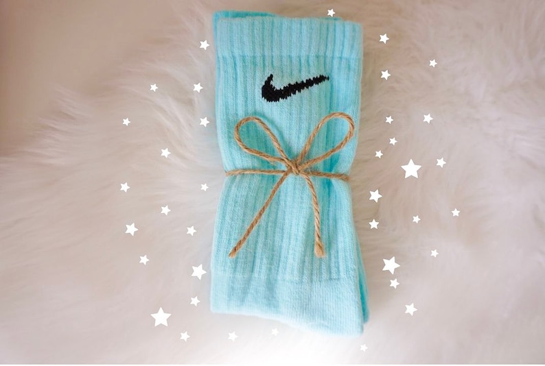 May include: A pair of light blue socks with the Nike swoosh logo in black. The socks are tied with a brown twine bow. The background is white with white star shapes.
