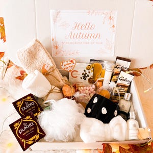 May include: A gift box containing two Pukka vanilla chai tea bags, a white bath pouf, a black mug with white ghost patterns, a white headband, beige fuzzy socks, Galaxy hot chocolate packets, mini marshmallows, and small skincare bottles.