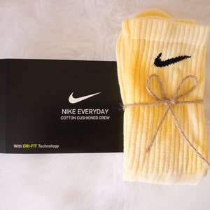 May include: A pair of light yellow Nike Everyday cotton cushioned crew socks with a black Nike swoosh logo, tied with twine, next to a black box with the Nike logo and text.