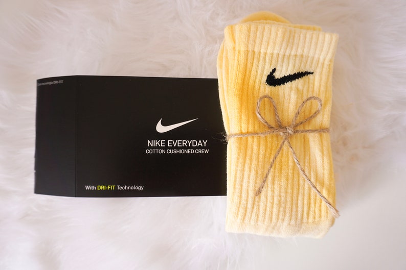 May include: A pair of light yellow Nike Everyday cotton cushioned crew socks with a black Nike swoosh logo, tied with twine, next to a black box with the Nike logo and text.