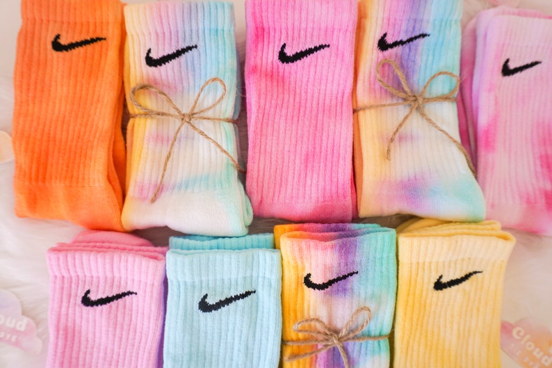 May include: A collection of colorful tie-dye socks with the Nike swoosh logo in black. The socks are in various shades of orange, pink, blue, and yellow. Some pairs are tied together with twine. The socks are ribbed and appear to be made of a soft material.