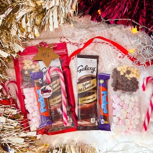 May include: A festive gift set featuring instant hot chocolate, fudge bars, marshmallows, and candy canes. The set includes clear bags filled with treats, a star-shaped tag, and is surrounded by gold and red tinsel.
