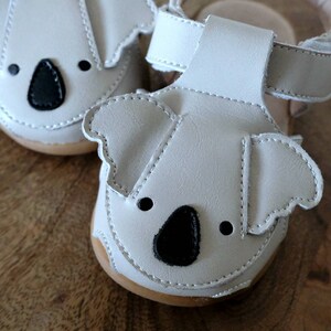 May include: A pair of off-white baby sandals with koala bear faces. The sandals have black noses and eyes, and a strap. The soles are light brown. The sandals are made of a soft material.