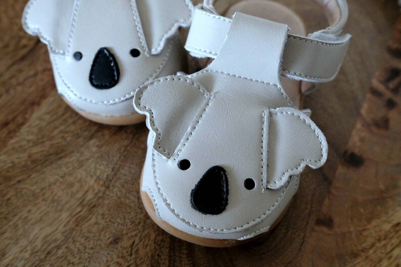 May include: A pair of off-white baby sandals with koala bear faces. The sandals have black noses and eyes, and a strap. The soles are light brown. The sandals are made of a soft material.