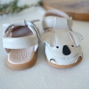 May include: A pair of off-white baby sandals. One sandal features a koala face design with black accents. Both sandals have light brown soles and adjustable straps. The brand name "FRANC DANI" is visible on the insole.