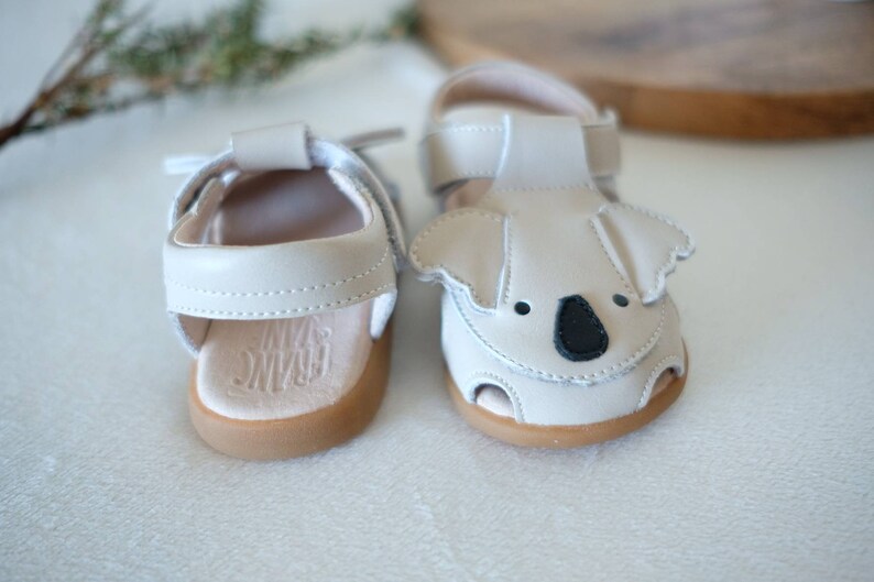 May include: A pair of off-white baby sandals. One sandal features a koala face design with black accents. Both sandals have light brown soles and adjustable straps. The brand name "FRANC DANI" is visible on the insole.