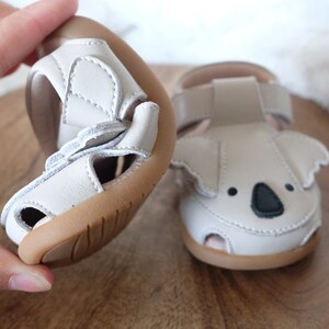 May include: Light gray baby sandals with a koala face design. The sandals have a flexible sole and adjustable straps. The koala face features black eyes and a black nose. The sandals are made of a soft material and are designed for comfort.